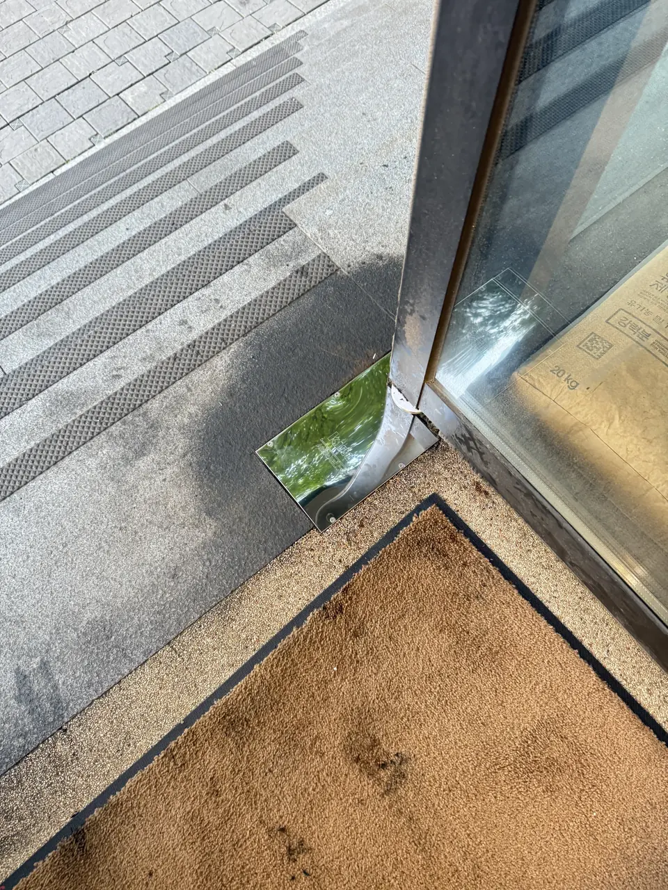 현관 매트 옆 강화유리문 하단 힌지와 바닥 접합부